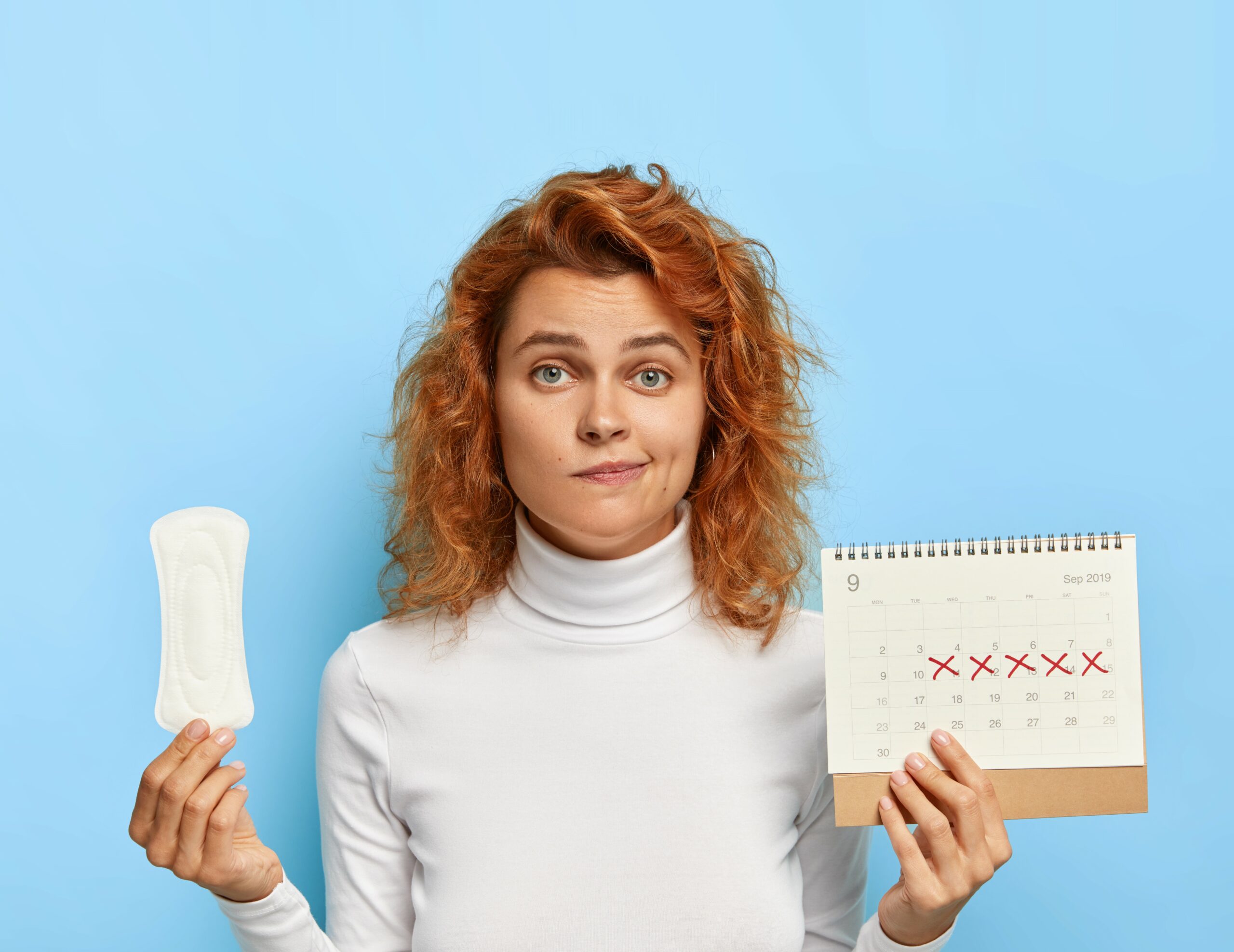 woman holds sanitary napkin and menstruation calendar with marked red days, suffers from period cramps, controls her women health, has periods this week, poses over blue background